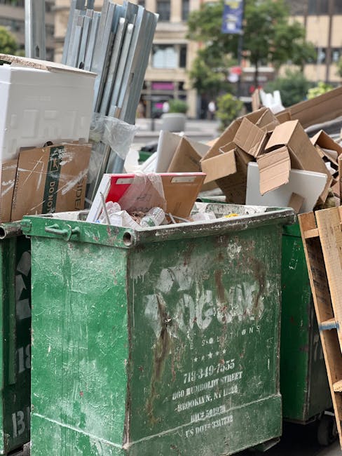 A large, weathered green wheelie bin with visible rust and dirt stains, positioned outdoors on a paved surface, filled with mixed waste including crumpled paper, plastic bottles, and broken cardboard boxes. Surrounding the bin are several flattened cardboard boxes, some leaning against the bin, and others stacked nearby. To the left, metal poles or fixtures are partially visible, wrapped in clear plastic. In the background, a city street scene can be seen, featuring blurred storefronts, parked cars, and trees, indicating a busy urban environment. The scene appears to be part of an onsite rubbish collection or disposal process, possibly organized by House Clearance Dulwich, highlighting a typical rubbish removal task involving the collection of various waste materials. The lighting suggests natural daylight, with no significant shadows or reflections, emphasizing the practical and neutral depiction of waste management activities in a public or semi-private setting.
