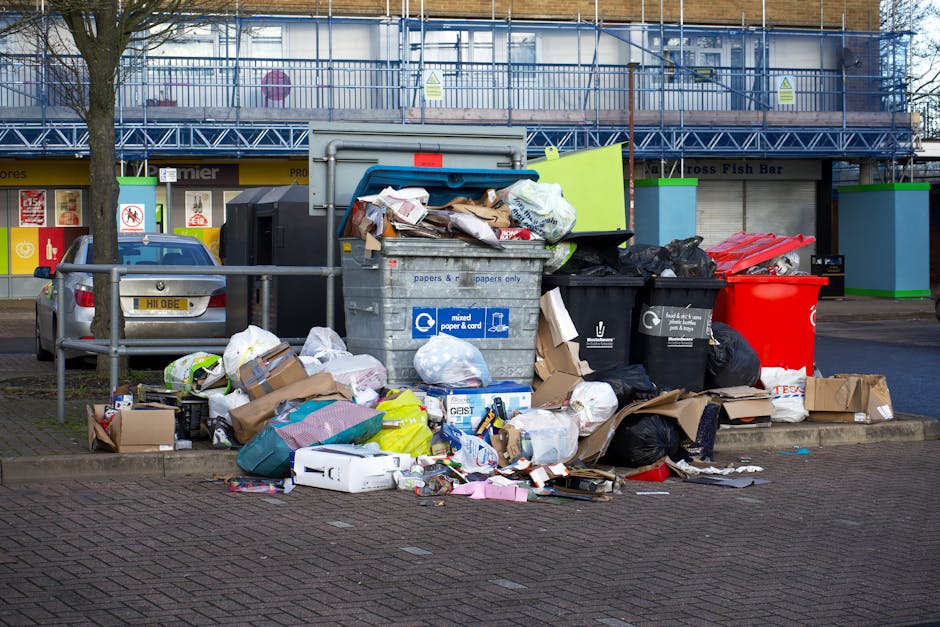 A collection of overflowing waste and recycling bins situated on a paved area adjacent to a parking space, with various types of rubbish including black and black-and-clear plastic bags, flattened cardboard boxes, paper waste, and scattered packaging materials spilling onto the ground. The central grey bin labeled for mixed paper and card is partially open, revealing crumpled paper and cardboard inside. To the right, a red recycling bin, a black bin, and another black bin are visible, all filled with trash. In the background, a building under construction is covered with blue scaffolding and safety netting, with storefronts and signage below indicating a commercial area. A grey vehicle is parked on the left, partially obscured by a metal fence separating the waste area from the sidewalk. The scene is daytime with diffuse natural light, and the environment reflects an urban setting where waste accumulation suggests the need for collection services, potentially provided by local or independent waste removal companies like House Clearance Dulwich.