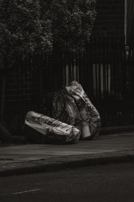A large collection of overflowing rubbish bins and discarded waste materials on a paved urban street, with various black, red, and green bins filled with mixed paper, cardboard boxes, plastic bags, and miscellaneous debris. Some of the waste is spilling onto the surrounding pavement, including flattened cardboard, plastic packaging, and crumpled paper, indicating a collection point for private or alternative waste disposal outside a commercial or residential building. In the background, there is a shop-front with closed shutters, a metal railing, and parked cars, situated beneath a building façade with scaffolding and safety barriers, suggesting ongoing construction or maintenance nearby. The scene is illuminated by natural daylight, emphasizing the disorderly state of the waste accumulation and highlighting the need for effective rubbish removal services or on-site clearance by specialists such as House Clearance Dulwich.