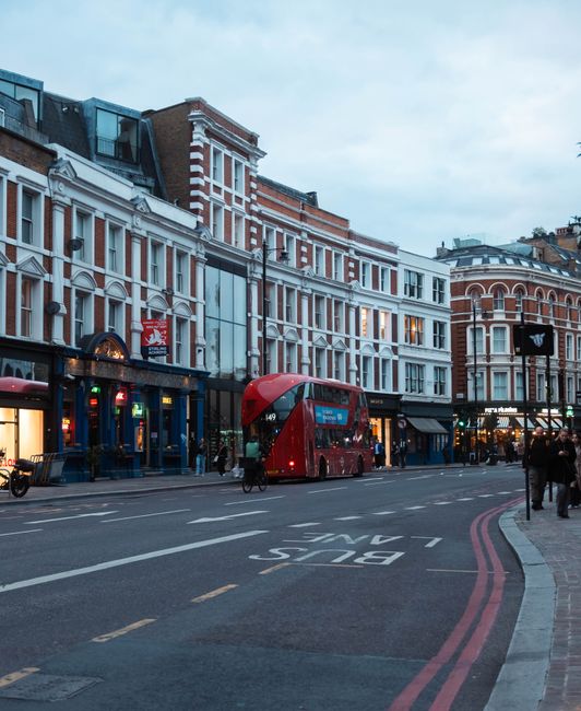 A busy urban street scene in Dulwich with a row of multi-story buildings featuring white and red brick facades, large windows, and decorative architectural details. The street is lined with shops, some illuminated with warm lighting, and a red double-decker bus is prominently seen in the center of the image, approaching the intersection. Pedestrians are walking along the sidewalk, and bicycles are parked near the shop entrances. The road has clear lane markings, including a bus lane marked with the words 'BUS LANE' in white paint. The environment appears to be late afternoon or early evening with a slightly overcast sky, and the ambient lighting highlights the textures of the brickwork and the reflective surfaces of the bus and shop windows. This scene exemplifies an active city street, demonstrating elements of urban transportation, retail activity, and pedestrian movement, which are areas where professional waste disposal, such as those offered by House Clearance Dulwich, might be involved in managing commercial rubbish from local shops and businesses.