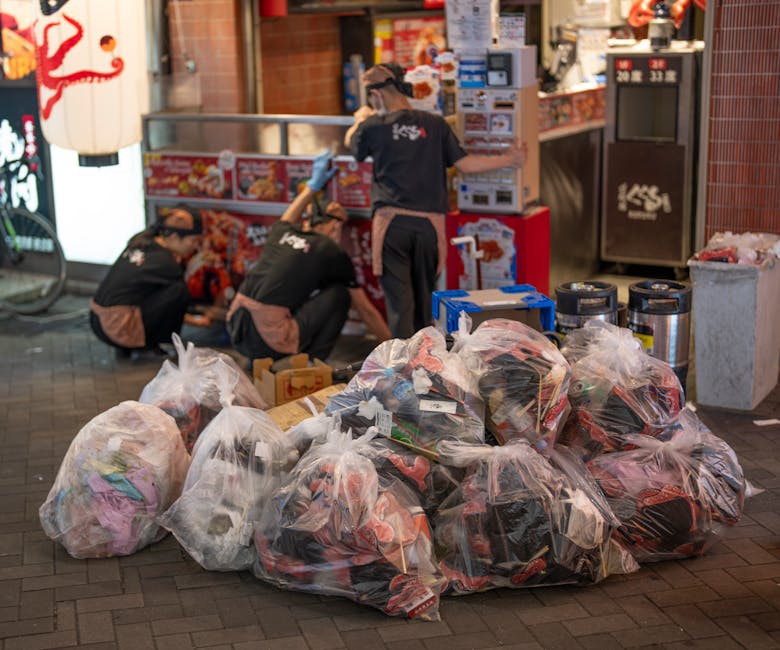 A collection of large transparent plastic bags filled with mixed household waste materials such as clothes, packaging, and other refuse, situated on a brick-paved sidewalk in front of a Japanese restaurant illuminated by street lighting. Behind the bags, three workers in black shirts with logos, gloves, and caps are engaged in sorting or lifting additional rubbish from a compartmentalized waste collection unit. The background features a brightly lit storefront with red-tiled walls, a vending machine, and various boxes and containers stacked nearby. The scene suggests an on-site rubbish clearance operation that may be part of a private waste removal service, aligned with the terms of independent rubbish collection and disposal, as managed by companies like House Clearance Dulwich. The environment appears typical of an urban commercial area at dusk, with the temporary accumulation of waste awaiting collection or transfer for disposal or recycling, demonstrating an organized approach to waste handling in accordance with professional rubbish removal practices.