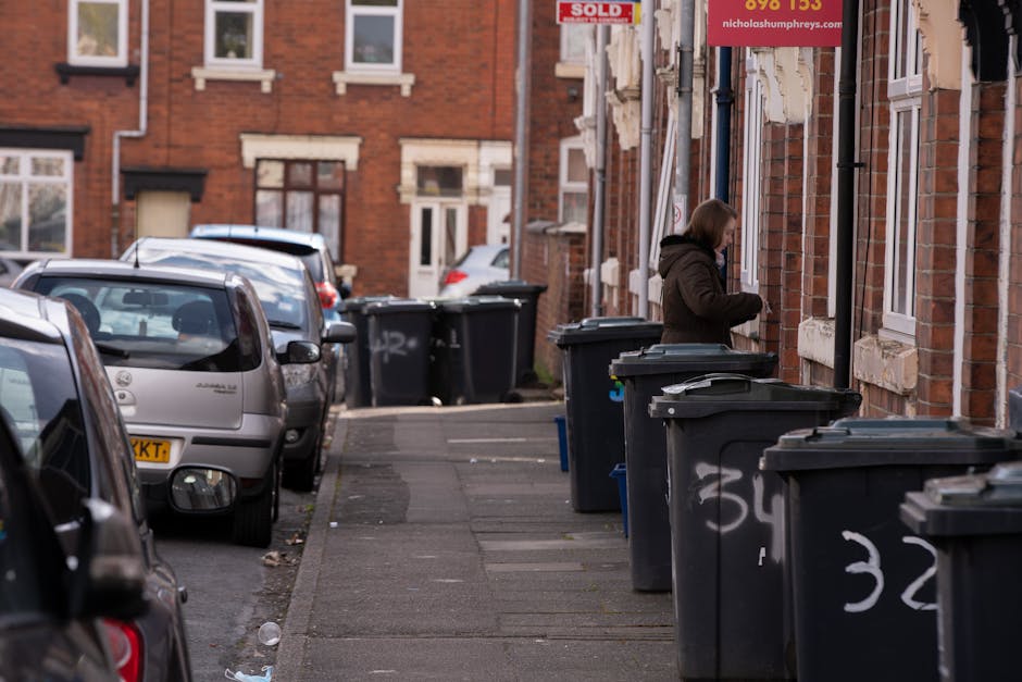A row of black wheelie bins with closed lids positioned along a residential pavement in front of brick terraced houses. The bins are spaced at regular intervals, some with white chalk numbers and marks on their fronts. A woman dressed in a dark coat is standing next to one of the bins, appearing to access or manage waste. The background features parked cars lining the street, including a silver vehicle in the foreground and several others further down the road. The brick buildings behind the bins have white-framed windows, and some houses have small signs or advertisements posted on the walls, with one visible sign indicating the property has been sold. The scene is illuminated by natural daylight, contributing a neutral tone that highlights the practical setting of on-site waste management as part of independent rubbish collection services in an urban residential area, aligning with the context of alternative waste handling options for Dulwich homes. The environment looks clean but informs of regular waste disposal activity typical of private rubbish removal services.